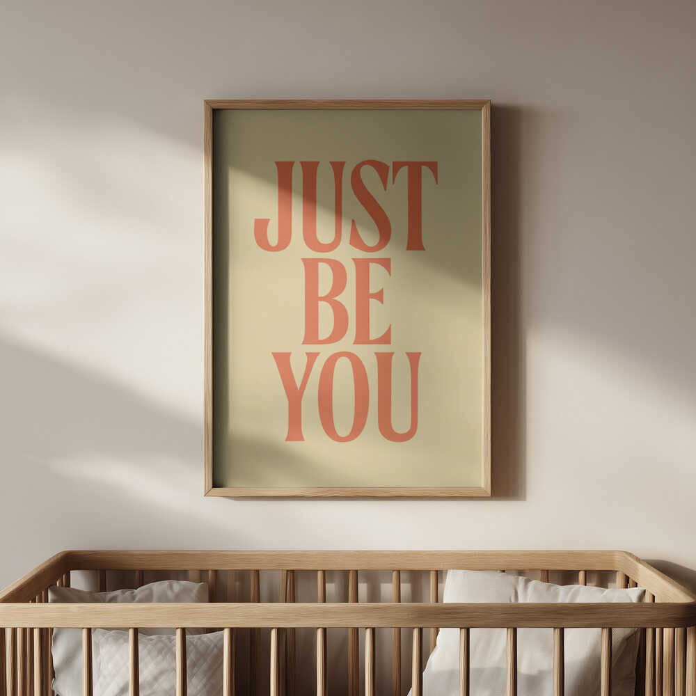 Framed nursery poster with 'JUST BE YOU' text in orange on a light yellow background hanging above the baby crib